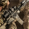 Widok zza kolby karabinu na zamontowaną lunetę Vortex AMG 1-10x24 FFP. Skoncentrowane ujęcie z perspektywy strzelca celującego w daleki obiekt na otwartej przestrzeni, prezentujące optykę HD klasy premium i 34 mm tubus w pełnej gotowości operacyjnej.